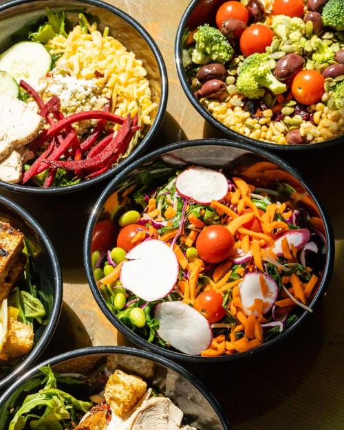 A variety of colorful salad bowls with greens, vegetables, grains, and proteins arranged on a wooden surface.