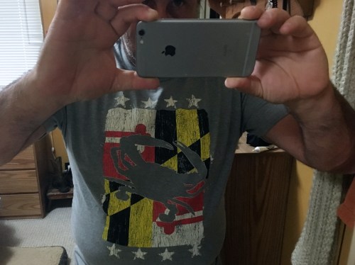 A person taking a selfie in a gray t-shirt featuring a scorpion design and a colorful Maryland flag background.