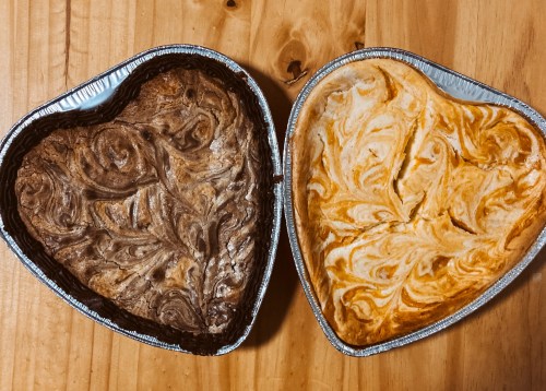 Two heart-shaped desserts in aluminum pans: one chocolate marble and one orange swirl, placed on a wooden surface.