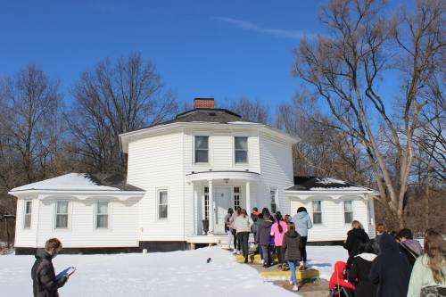 A group of people walks towards a large white house on a snowy day, surrounded by bare trees and a clear blue sky.