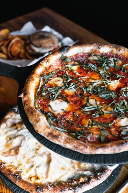 A close-up of two pizzas on a wooden serving board, topped with fresh basil and cheese, with snacks in the background.