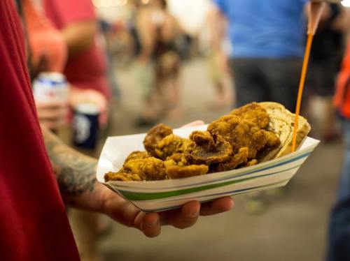 A person holds a tray of fried chicken with bread, surrounded by a bustling food festival atmosphere.