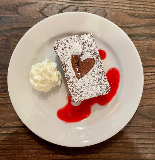 A chocolate dessert with a heart shape, dusted with powdered sugar, served with whipped cream and raspberry sauce.