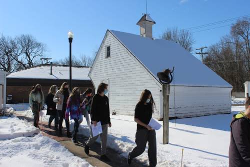 A group of people wearing masks walks along a snowy path beside a white building on a sunny day.