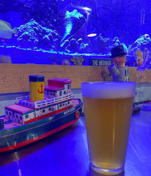 A glass of beer sits on a table next to a toy boat and a colorful aquarium in the background.