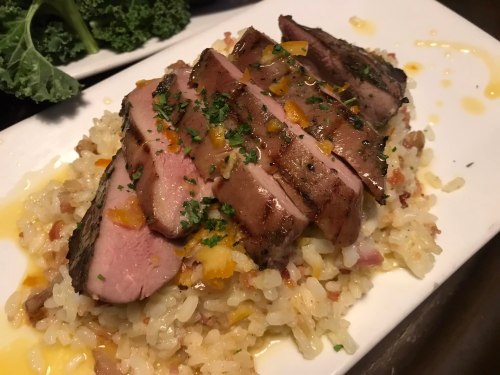 Sliced grilled meat on a bed of rice, garnished with herbs and orange zest, served on a white plate.
