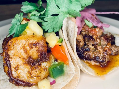 Two tacos on a plate: one with shrimp and pineapple salsa, the other with seasoned beef, topped with cilantro and pickled onions.