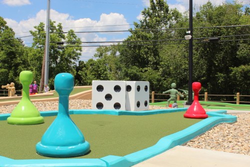 Colorful game pieces and a large dice on a mini-golf course surrounded by greenery and blue skies.