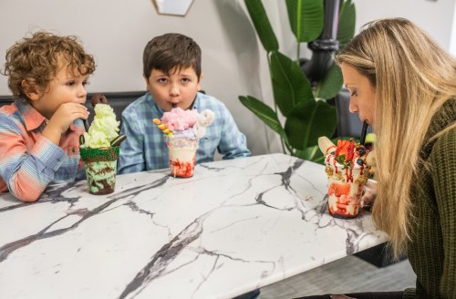 Two children and an adult enjoy colorful ice cream sundaes at a marble table, smiling and sharing a fun moment.