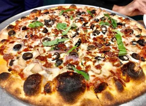 A close-up of a pizza topped with olives, bacon, onions, and fresh basil on a golden crust.