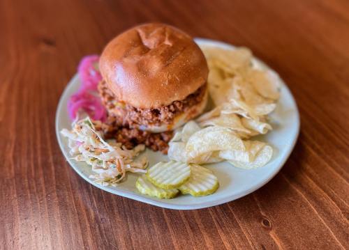 A plate with a messy sandwich, pickles, coleslaw, and potato chips on a wooden table.