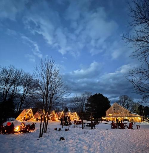 A winter scene with cozy cabins, snow-covered ground, and a glowing fire pit under a cloudy evening sky.