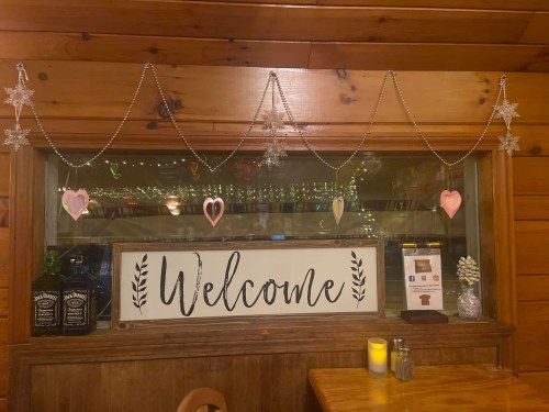 A cozy wooden interior with a "Welcome" sign, decorated with hearts and snowflakes, and a candle on the table.