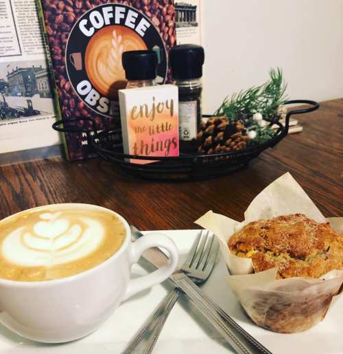 A cozy coffee setup featuring a latte with latte art, a muffin, and a sign that says "enjoy the little things."