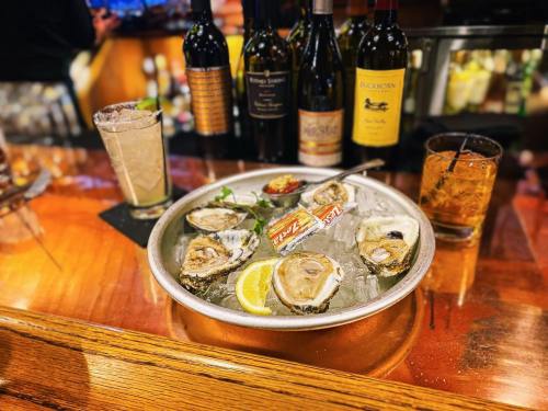 A platter of fresh oysters on ice, garnished with lemon, alongside cocktails and bottles of wine in the background.