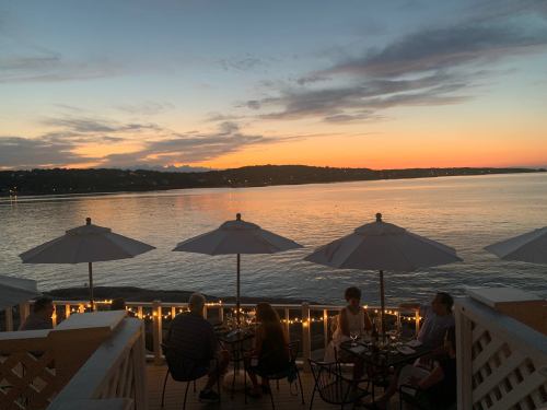 A serene sunset over a calm lake, with diners under umbrellas on a deck, surrounded by soft lighting.