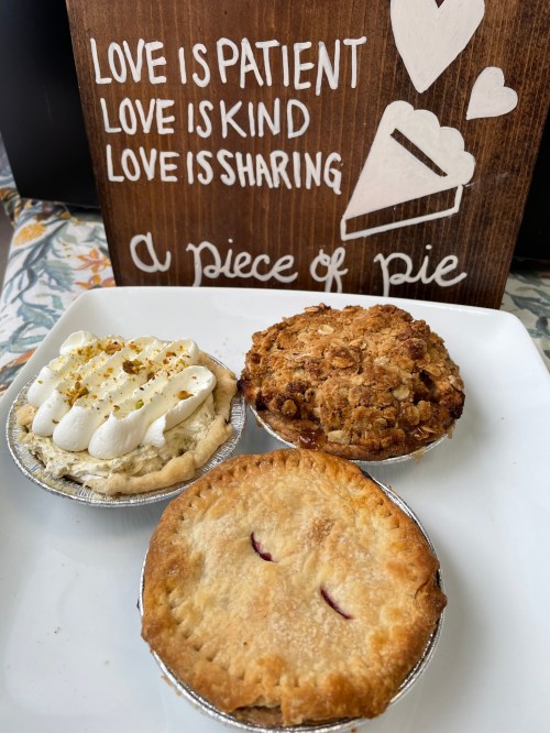 Three delicious pies on a plate: a cream pie, a fruit crumble, and a double-crust pie, with a wooden sign in the background.