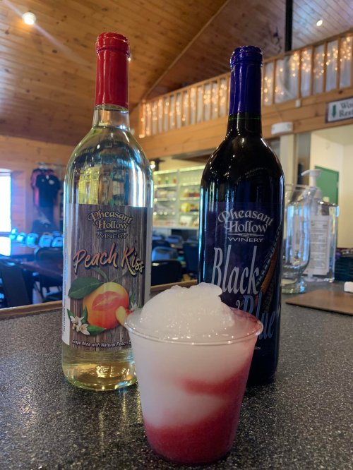 Two wine bottles labeled "Peach Kiss" and "Black & Blue" beside a frosty drink in a cup with a layered red and white appearance.
