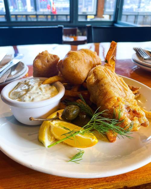 A plate of crispy fish and chips with tartar sauce, lemon wedges, and pickles on a wooden table.