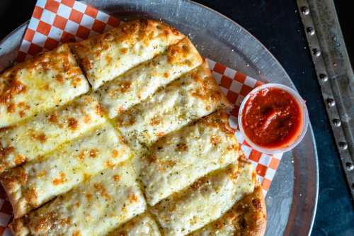 A tray of cheesy garlic breadsticks with marinara sauce on the side, served on a silver platter with a checkered paper.