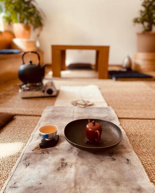 A serene tea setup with a teapot, cup, and plate on a woven mat, surrounded by plants and wooden furniture.