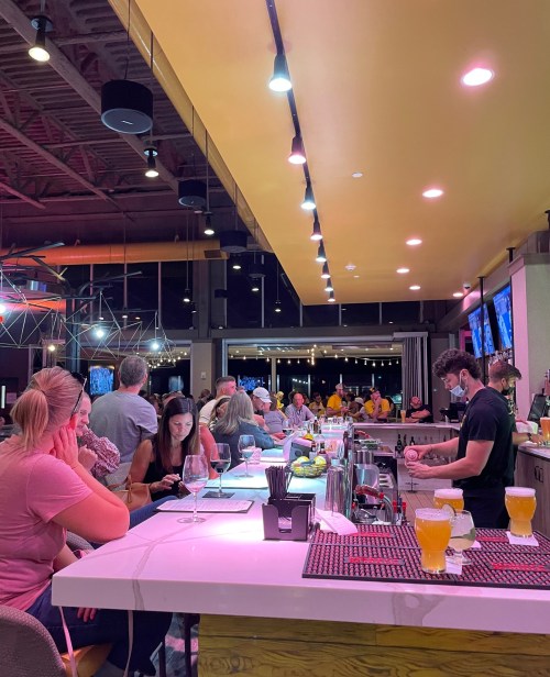 A lively bar scene with patrons enjoying drinks, a bartender preparing cocktails, and bright yellow lighting overhead.
