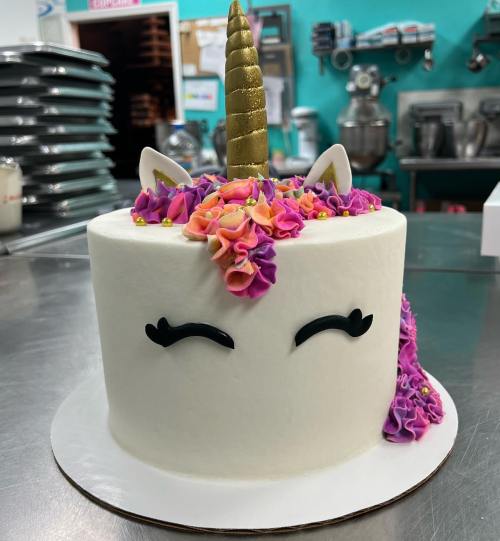 A whimsical unicorn cake with a golden horn, colorful frosting flowers, and closed eyes, set in a bakery.