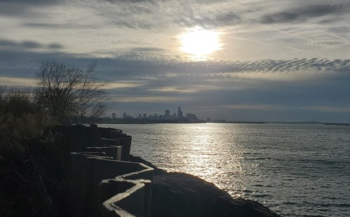 A serene waterfront view with a city skyline under a cloudy sky, reflecting sunlight on the water's surface.