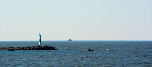 A calm sea with a distant boat and a lighthouse on a rocky jetty under a clear blue sky.
