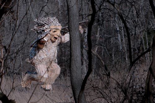 A large wooden sculpture of a troll-like figure leaps through a forested area, surrounded by trees and underbrush.
