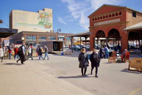 A bustling market scene with people walking, vendors, and a large mural on a building in the background.