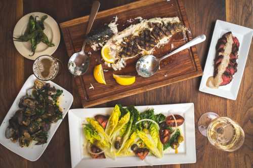 A wooden table with a variety of dishes: grilled fish, salad, clams, and a meat dish, accompanied by drinks.