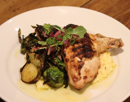 Grilled chicken thigh served with creamy polenta and sautéed Brussels sprouts, garnished with fresh herbs.
