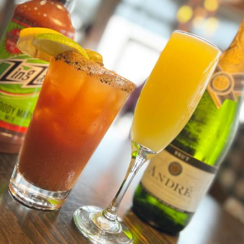 A vibrant cocktail setup featuring a Bloody Mary with a lime garnish and a mimosa beside a bottle of champagne.