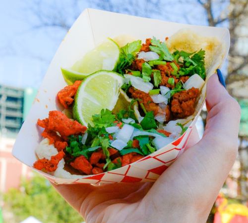A hand holding a taco filled with seasoned meat, onions, cilantro, and lime wedges in a paper tray.