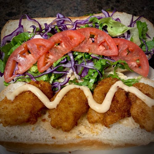 A sandwich with crispy fried shrimp, lettuce, tomato slices, and a drizzle of sauce on a soft white bread.