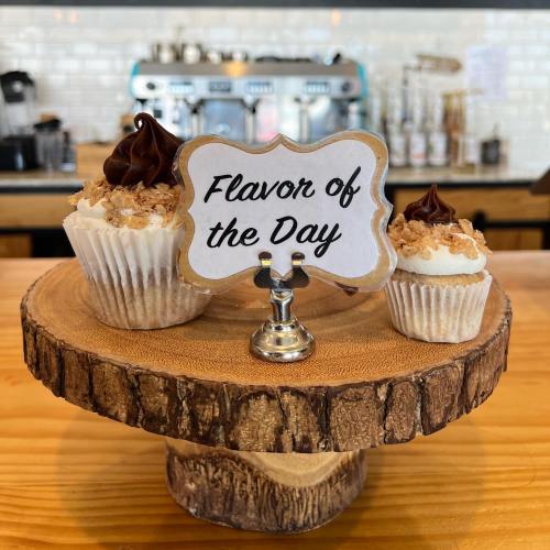 A wooden display stand holds two cupcakes topped with frosting and a sign reading "Flavor of the Day."
