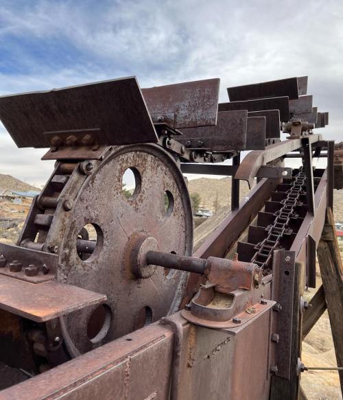 Close-up of a rusty industrial machine with gears and a conveyor belt against a cloudy sky.
