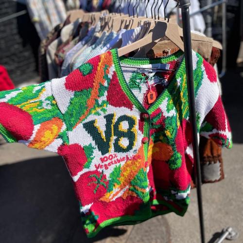 Colorful short-sleeve sweater featuring a vibrant vegetable print and "V8 100% Vegetable" text, displayed on a rack.
