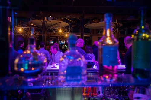 A vibrant bar scene with blurred bottles in the foreground and patrons socializing in the background under colorful lights.