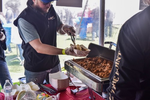 A person serving pulled pork from a catering tray at an outdoor event, with food containers and drinks on the table.