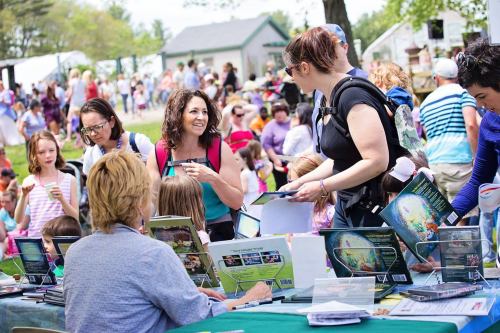 A busy outdoor event with people browsing tables, chatting, and enjoying activities in a lively atmosphere.