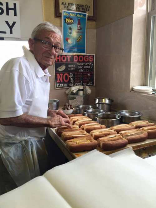 An elderly man prepares hot dogs at a counter, surrounded by trays and signs in a small food establishment.