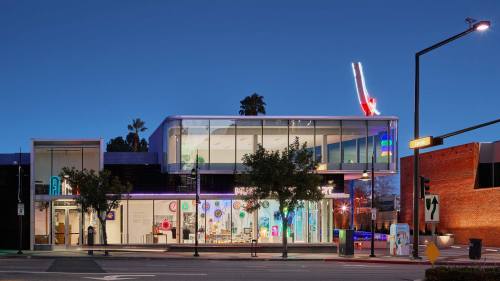 Modern glass building at dusk, featuring colorful lights and palm trees, with a vibrant urban atmosphere.