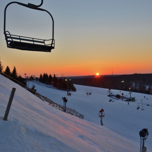 A ski lift chair hangs over a snowy slope at sunset, with silhouettes of trees and ski runs in the background.