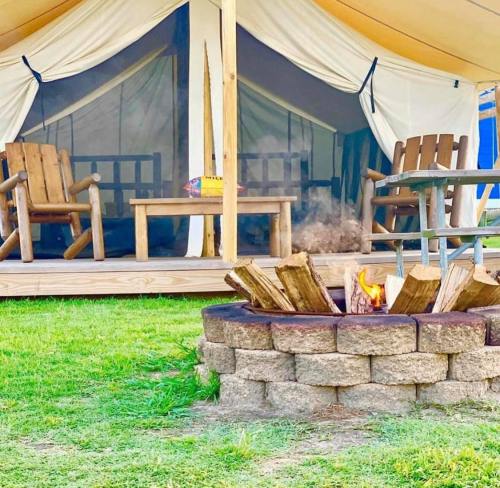 A cozy glamping tent with wooden chairs and a fire pit in front, surrounded by green grass.