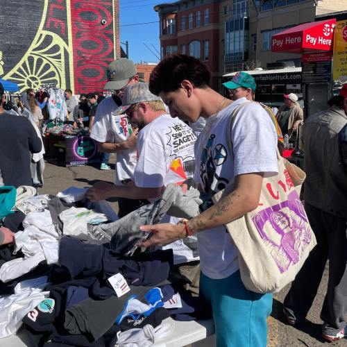 A person examines clothing at a bustling outdoor market, surrounded by others browsing through various items.