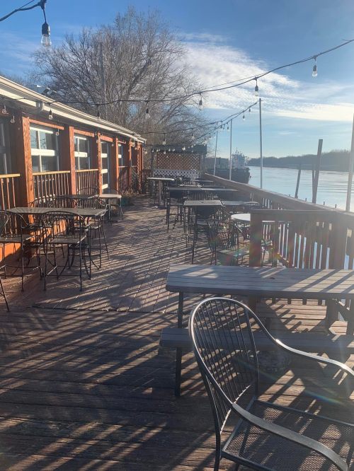 Outdoor dining area with tables and chairs by a river, surrounded by trees and string lights under a clear blue sky.