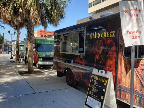 Food trucks parked on a city street, featuring "Fried's Mac & Cheez" and another truck in the background. Palm trees nearby.