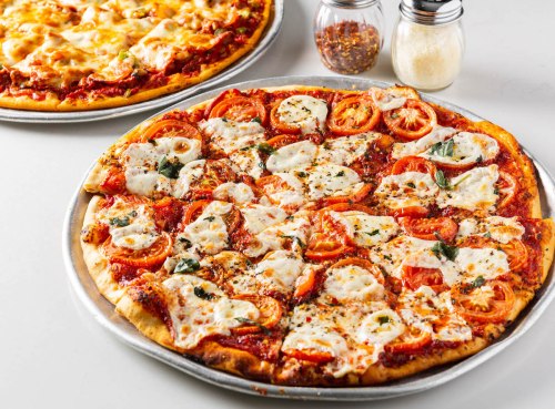 Two pizzas on a table; one topped with mozzarella, tomatoes, and herbs, the other with melted cheese. Seasoning jars nearby.